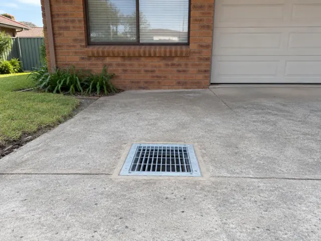 Concrete driveway with a square metal drain grate beside a brick house in Blacktown