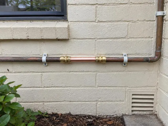 Repaired copper water pipe on a cream brick exterior wall with clamps and window trim in Woodcroft